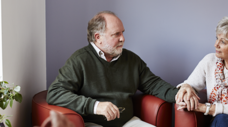 A man sat on a chair looking concerned, holding a ladies hand