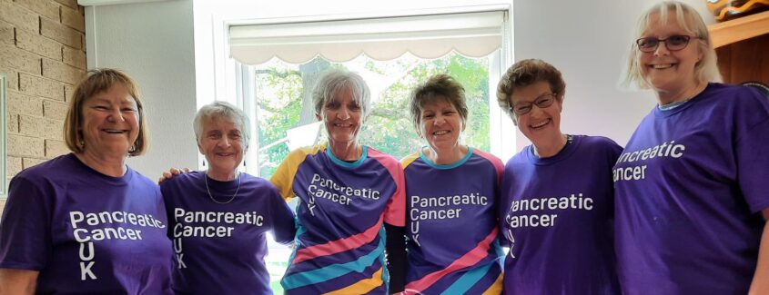 A group of women in purple t-shirts stood around a table, filled with cakes