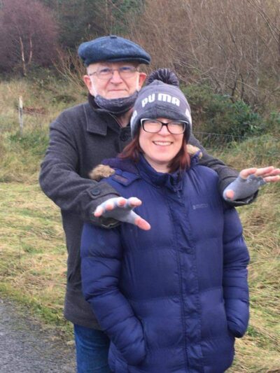 An older man leans on the shoulders of a younger woman. Both are wrapped up for a winter walk and smiling for the camera.