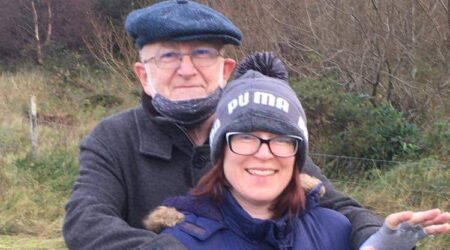 An older man leans on the shoulders of a younger woman. Both are wrapped up for a winter walk and smiling for the camera.