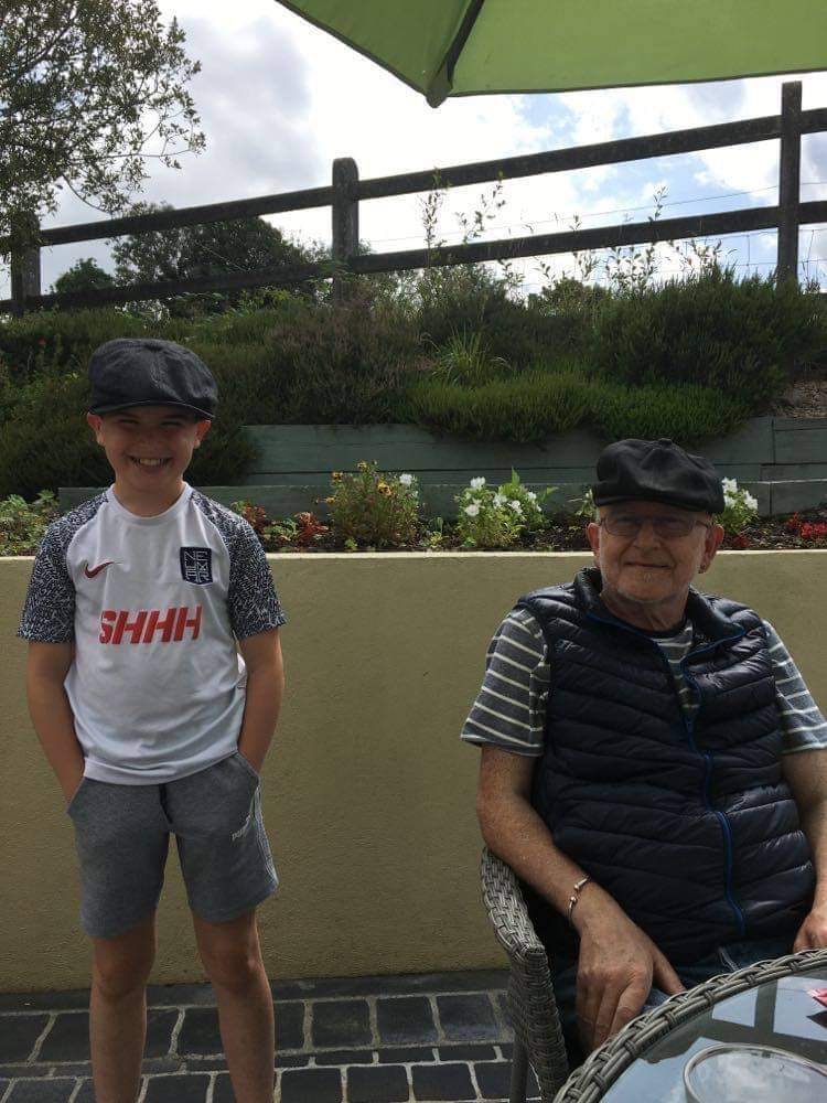 A young boy wearing a football shirt and a mature man both wear matching flat caps and smile on a sunny day.