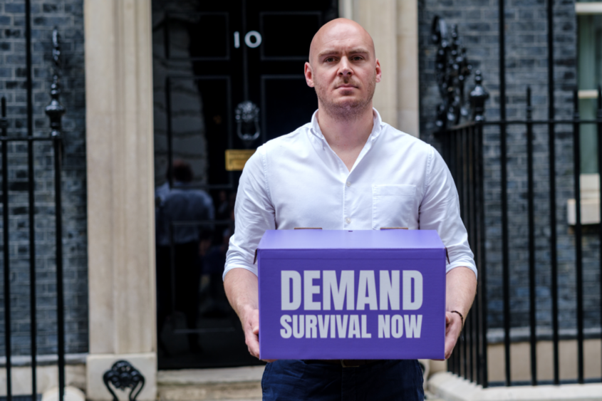 a man standing outside downing street holding a purple box that reads Demand survival now
