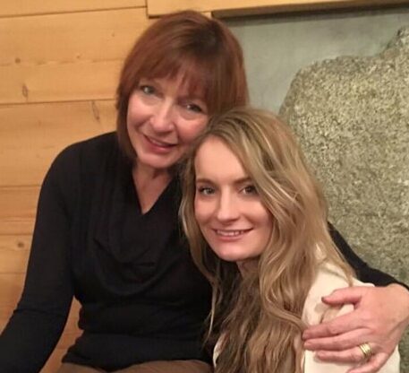 Woman sitting on a bench with her arm around her daughter, both smiling to camera