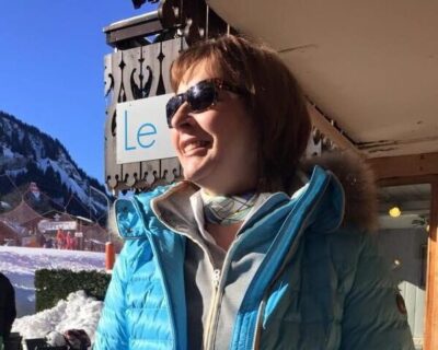 Woman standing in front of ski slopes wearing ski gear and shades and smiling