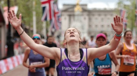 Woman running past the london marathon finish line with her arms in the air, with flags behind her