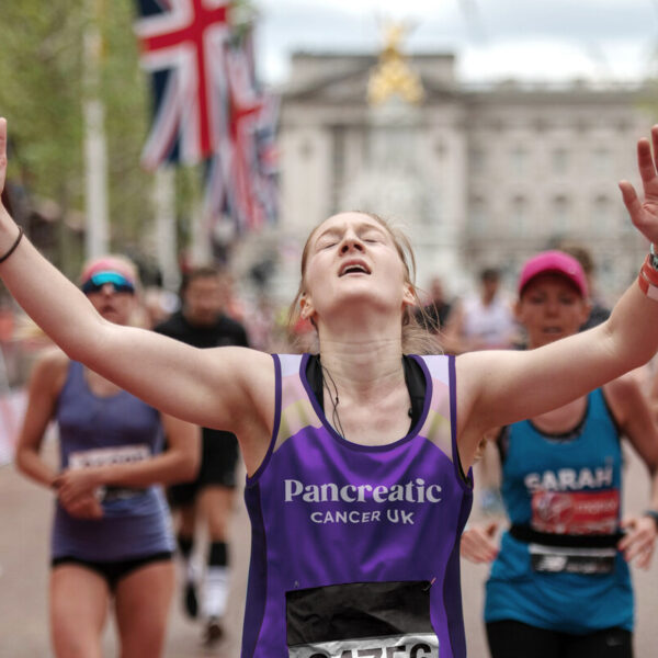 Woman running past the london marathon finish line with her arms in the air, with flags behind her