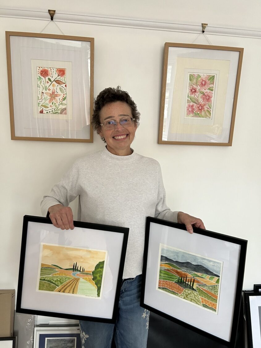 Woman holding two framed pictures and standing in front of two framed pictures on the wall, smiling to camera