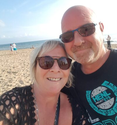 Man and wife selfie, both wearing shades and standing in front of beach