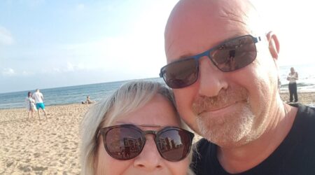 Man and wife selfie, both wearing shades and standing in front of beach