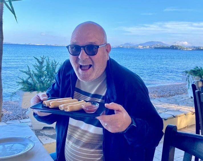 Man holding plate of food in an outside restaurant, sea and blue sky in the background