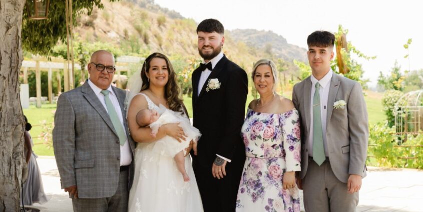 Family wedding photo with bride and groom, holding their baby and several other family members. Standing outside infront of countryside