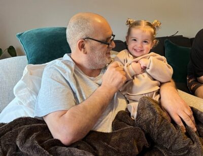 Elderly man sitting on sofa holding toddler age child, both snuggled under a blanket and smiling