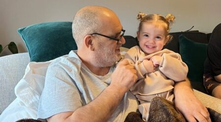 Elderly man sitting on sofa holding toddler age child, both snuggled under a blanket and smiling