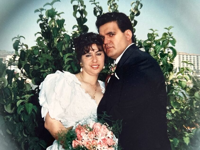 Older wedding photo of bride and groom. Standing in front of tree, holding each other and smiling to camera